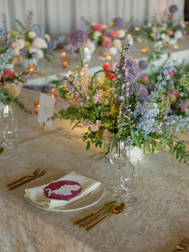 Low garden centerpiece of allium, delphinium, and stock on a patterned reception linen